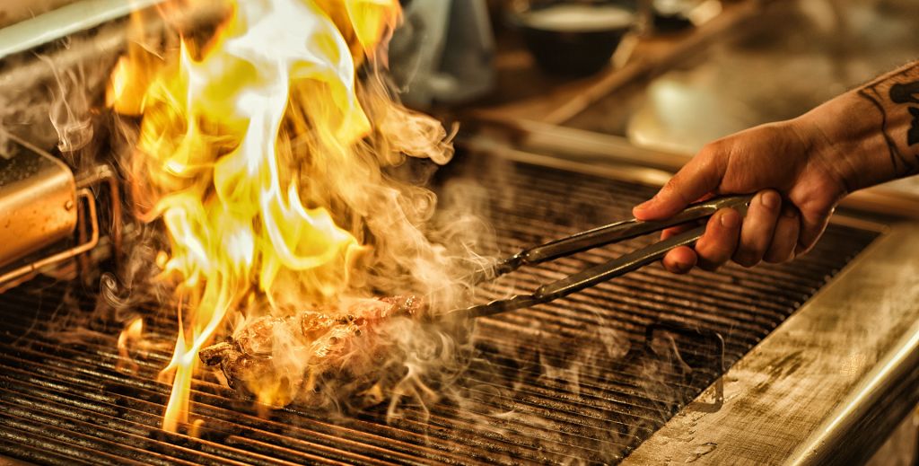 A steak is turned on a grill, flames bursting from the grate.